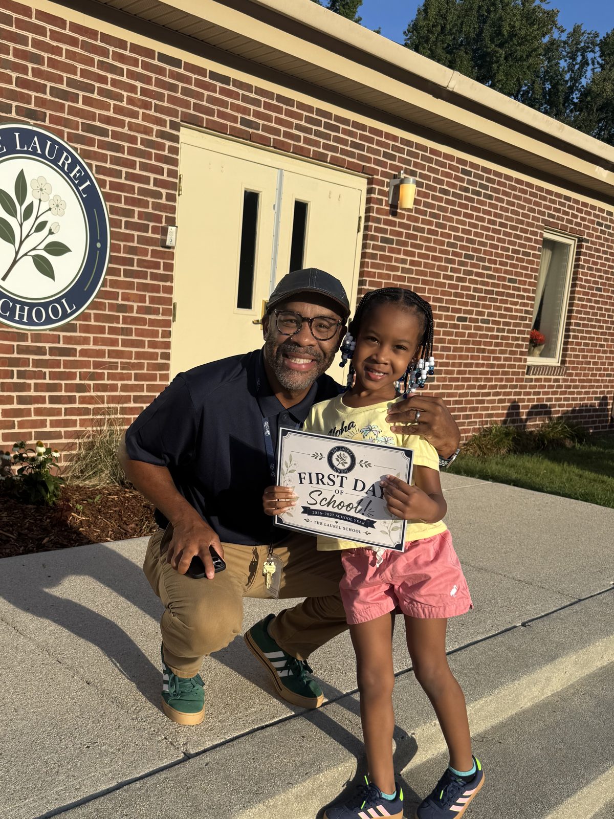 Dr. Harris kneeling beside a smiling Laurel student holding a First Day of School sign in front of the school